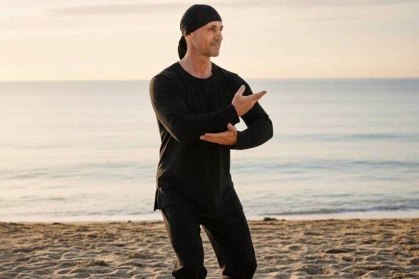 Una persona vestida de negro realizando una postura de artes marciales en una playa, con el océano al fondo. 3