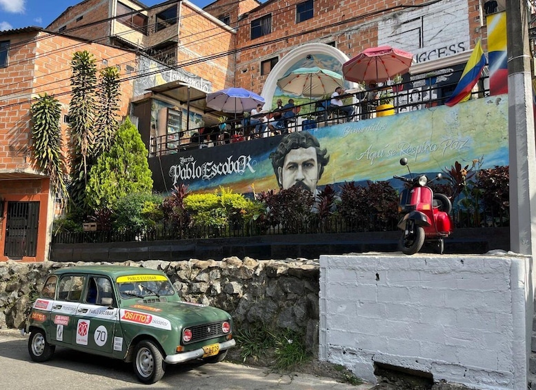 A vintage race car parked in front of a mural with a man's portrait and the text 'Pablo Escobar.' 1