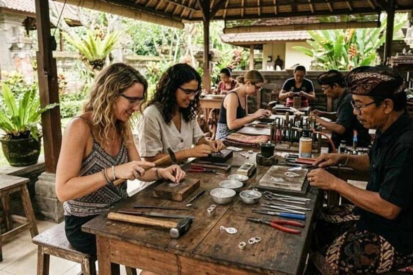 Personas dedicadas a una actividad artesanal, posiblemente la creación de bisutería, con herramientas y materiales sobre una mesa de madera en un entorno al aire libre con vegetación de fondo. 1