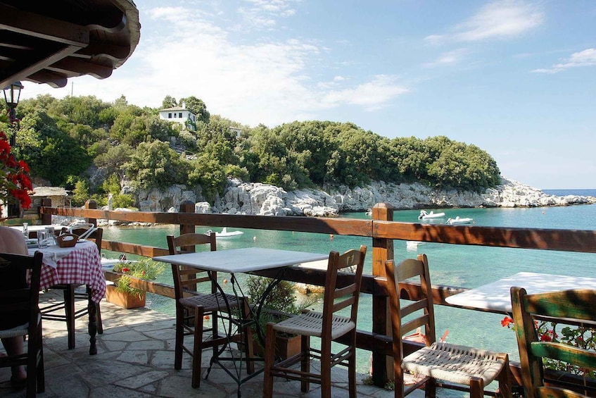 A seaside restaurant with outdoor seating overlooking a bay, rocky cliffs, and a white building on a hill. 3