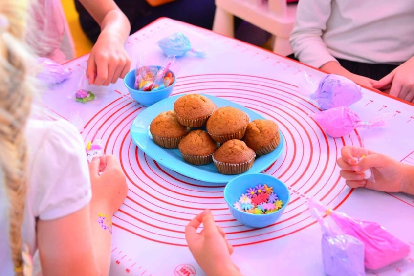 A group of people gathered around a table with a plate of muffins and a bowl of sprinkles. 3