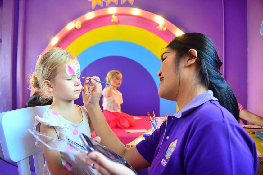 A child getting face paint applied by an adult in a brightly colored room with a rainbow and star decorations. 4
