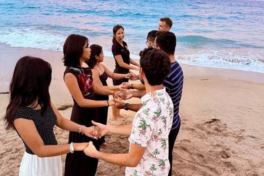A group of people holding hands in a circle on a beach with the ocean in the background. 2