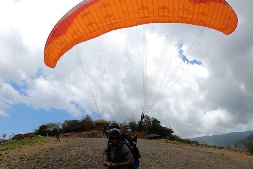 Iemand paraglidet onder een groot oranje zeil, met een heldere lucht en een glooiend landschap. 2