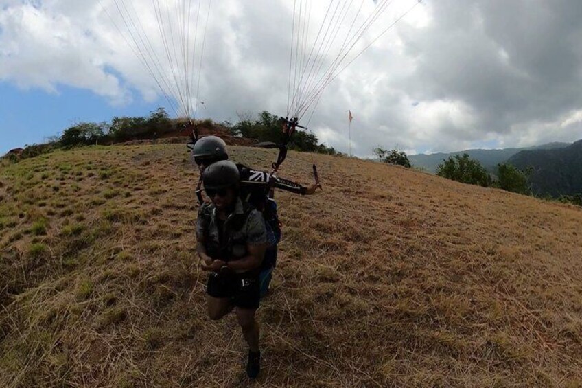 Een persoon met een parachuteharnas en helm, staand op een grasheuvel met een bergachtig achtergrond. 5