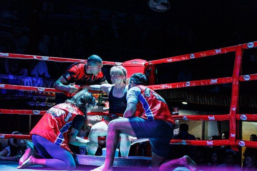 Ein Boxring mit einer Person in roten und blauen Sportklamotten, einer weiteren in Rot und einer dritten in Blau. 2
