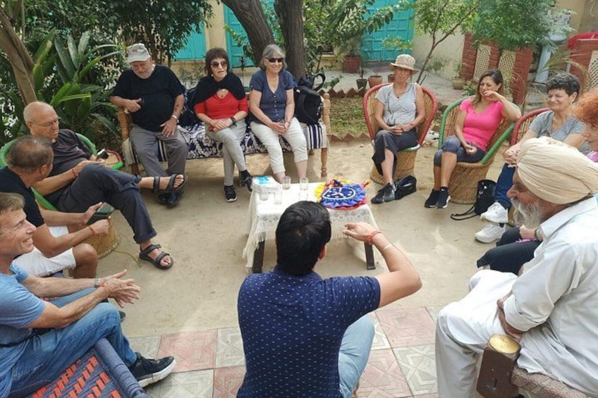 A group of people sitting in a circle outdoors, some on chairs, others on the ground, with a table in the center. 5