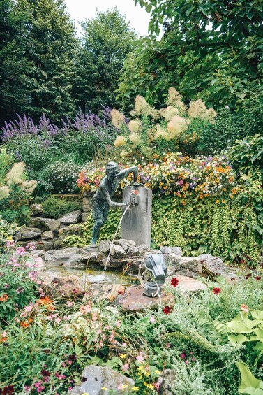 A garden statue of a figure with a hose, surrounded by a variety of colorful flowers and greenery. 2