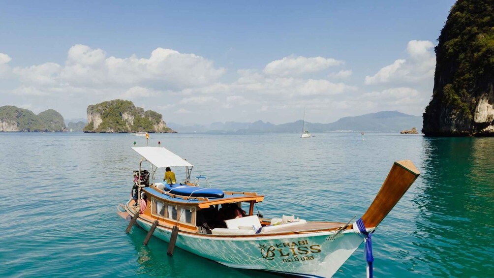 A traditional wooden boat named 'Krabi Bliss' docked at a calm sea with clear turquoise waters, surrounded by lush green islands and rocky cliffs under a partly cloudy sky. 2