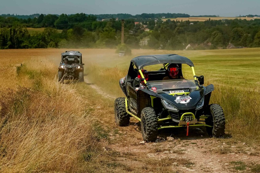 Two off-road vehicles on a dirt path in a rural area with fields and trees in the background. 1