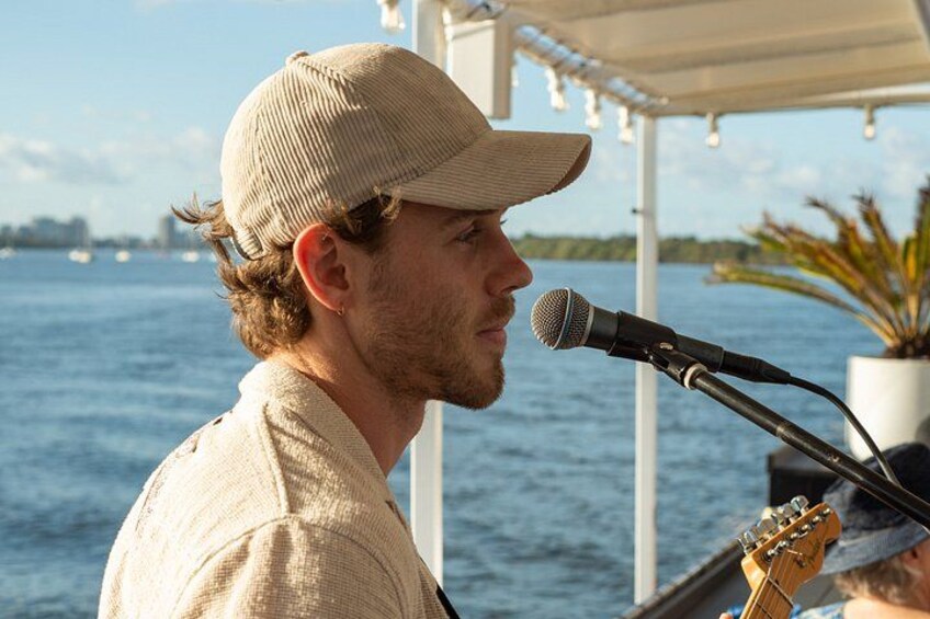A person playing a guitar on a boat with a microphone in front of them, water, and a city skyline in the background. 5