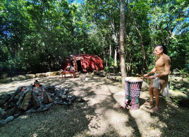 A person playing a drum in a forested area with a fire pit and a large red structure in the background. 4