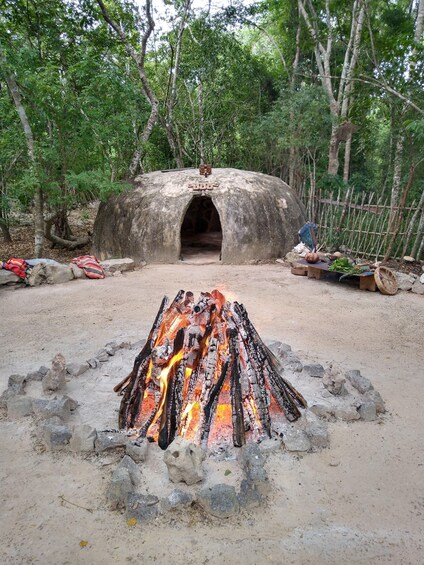 A traditional stone oven with an open entrance, surrounded by a forested area and a fire pit in the foreground. 2