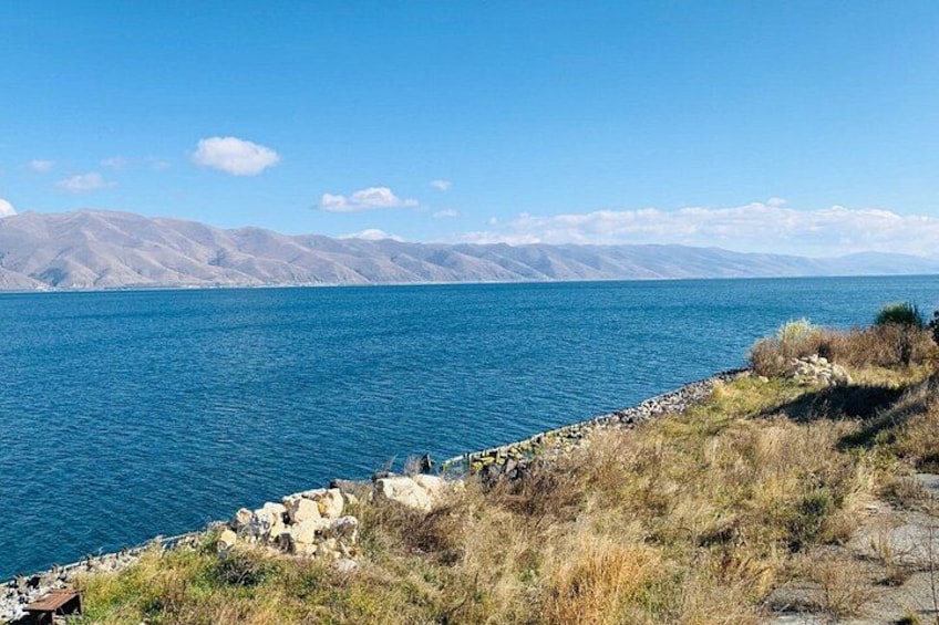 A lake with mountains in the background, a rocky shoreline, and sparse vegetation. 1