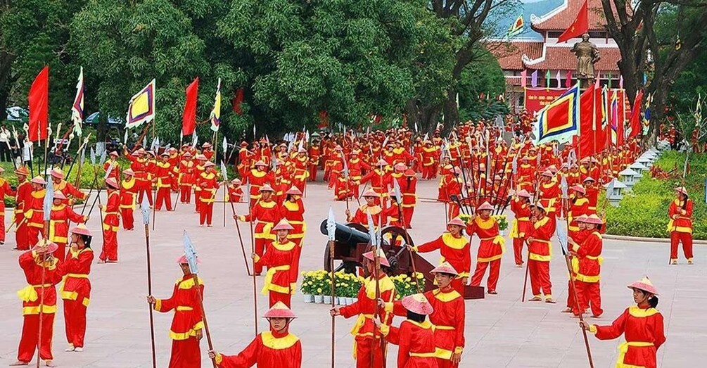 A large group of people in red uniforms with yellow trim, some carrying flags and others with a cannon, in a ceremonial formation. 3