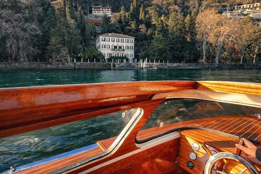 A wooden boat with a cabin, a steering wheel, and a view of a lake with a house in the background. 4
