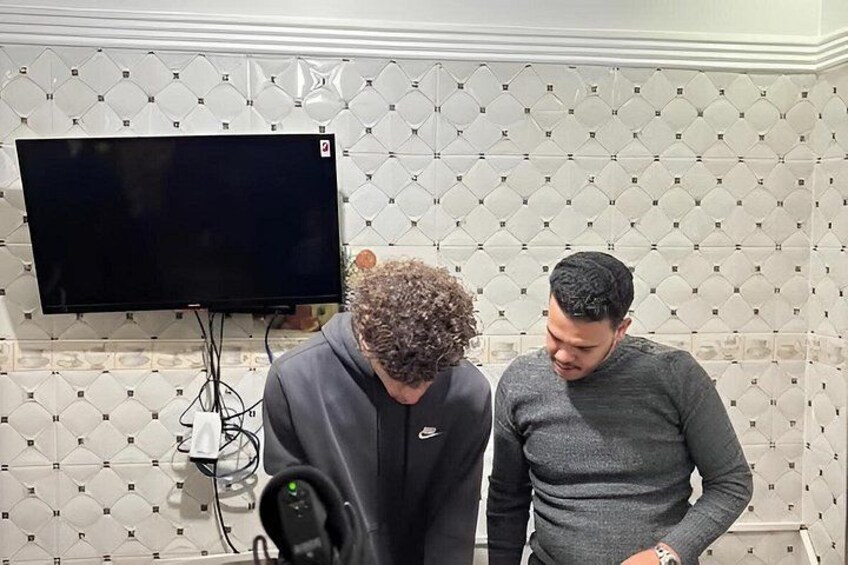 Two people in a room with a tiled wall and a flat-screen TV mounted above a hairdryer. 4