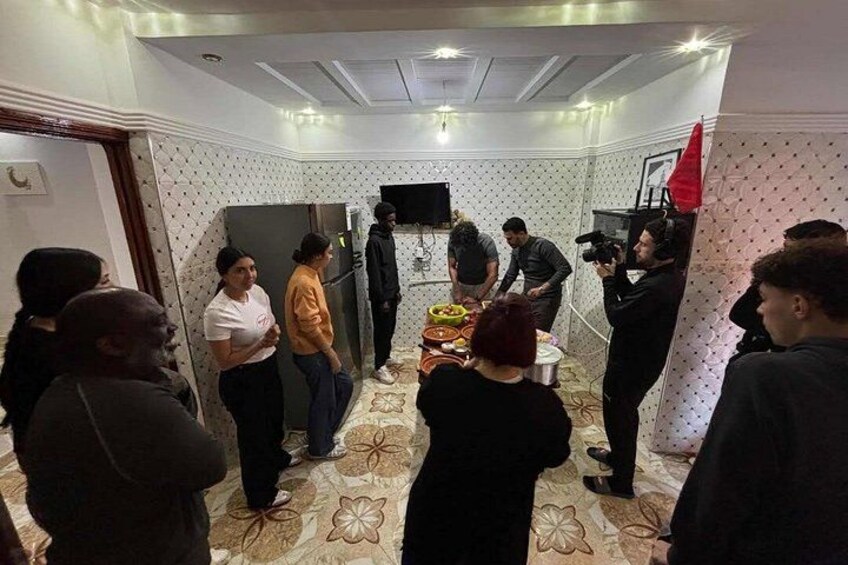 A group of people in a room with patterned walls and a tiled floor, some standing and some seated, with a camera and a red flag visible. 5