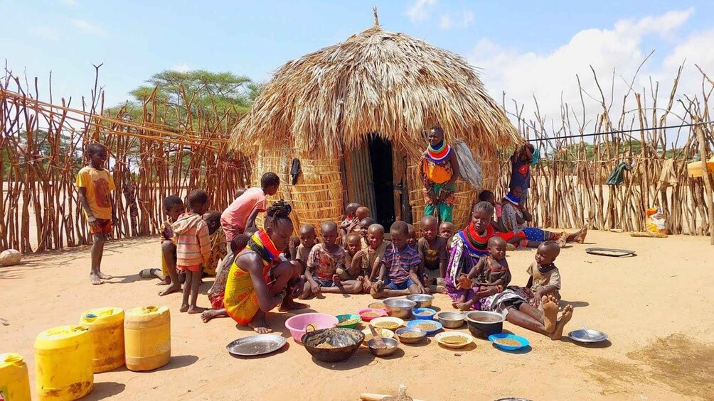 A rural community gathering around a thatched hut with children and adults seated on the ground, surrounded by various food containers. 1