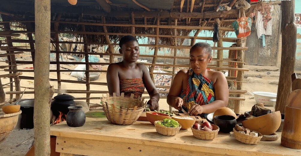 Two individuals at a wooden table with cooking utensils and baskets, likely preparing food in a rustic outdoor setting. 5