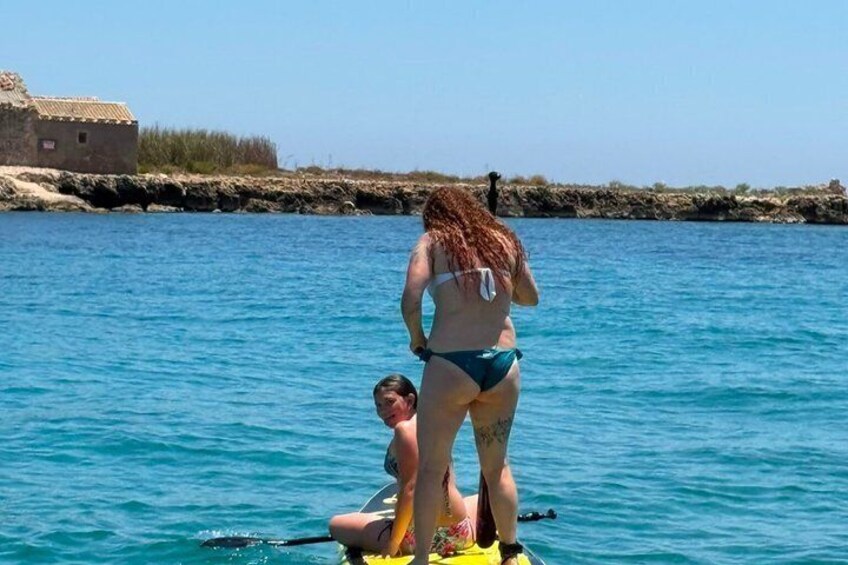 En person i bikini står på en gul surfbræt i vandet, en anden person sidder på brættet, og en klippefyldt kystlinje med en bygning i baggrunden. 5