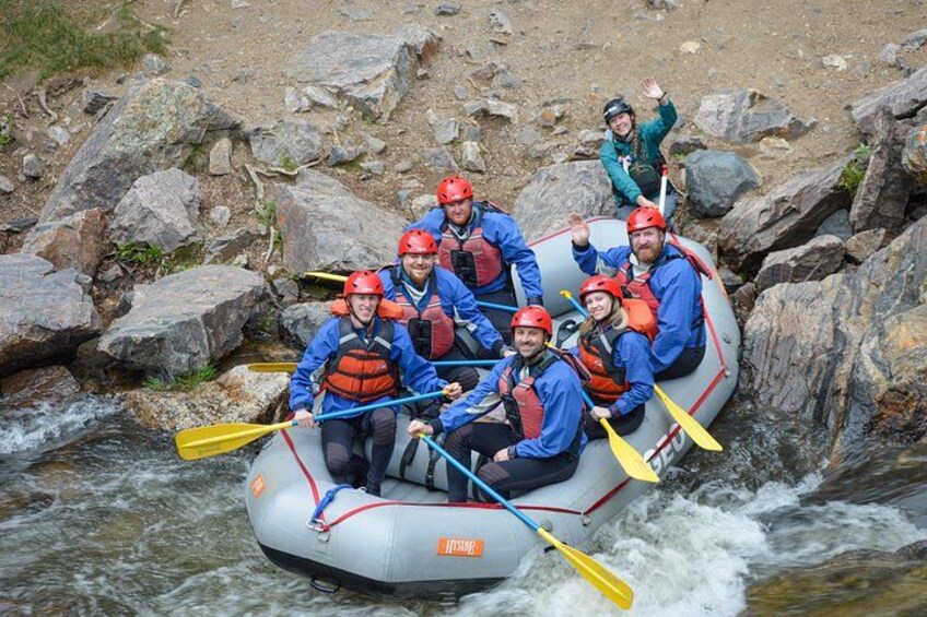 A group of people in a white inflatable raft navigating a river with rapids, wearing life jackets and helmets. 4