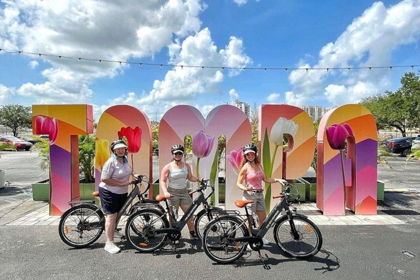 Three people standing in front of a colorful floral sign with bicycles. 2