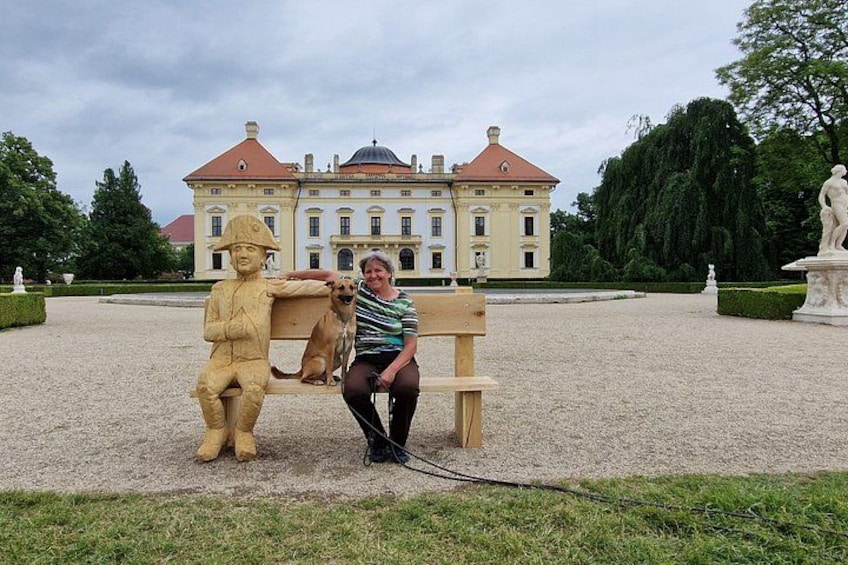 A person sitting on a bench with a dog, a statue, and a large building in the background. 3