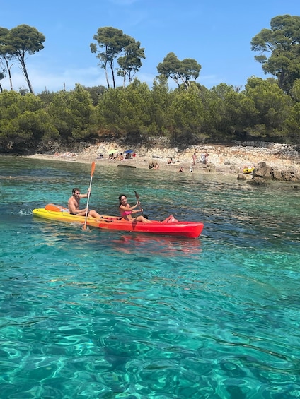 Two people kayaking on clear turquoise water with a rocky beach and trees in the background. 3