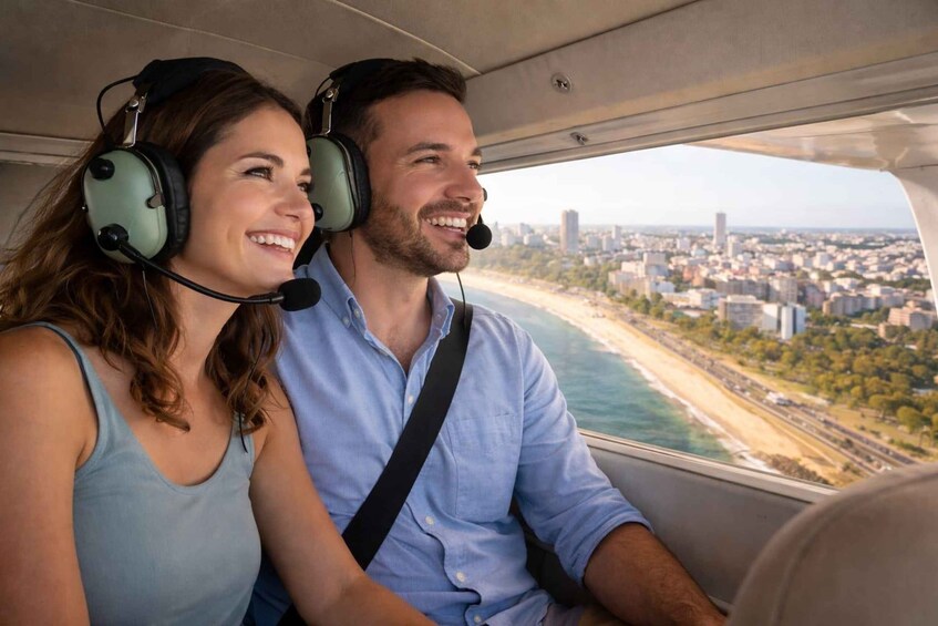 A person in a blue shirt and another in a light blue tank top, both wearing headphones, seated in a vehicle with a view of a coastal cityscape. 3