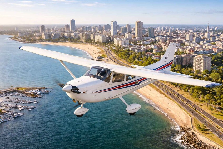 A small white airplane with red and blue stripes flying over a coastal city with a beach and marina. 1