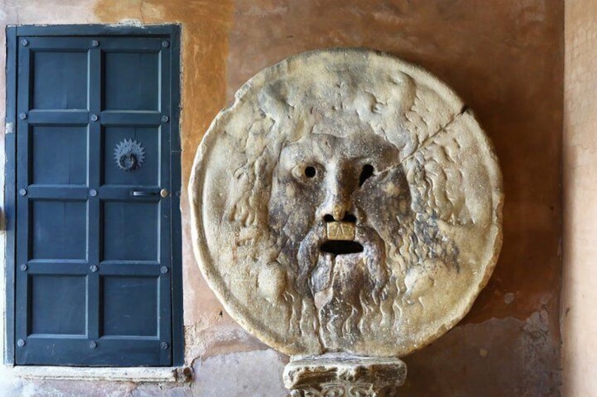 A weathered stone relief sculpture with a face, a dark door with a grid pattern, and a circular stone base. 2