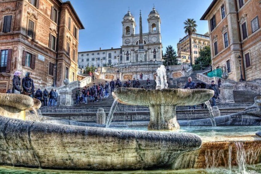 A historic fountain with multiple tiers and water spouts, surrounded by classical architecture and a prominent church in the background. 5