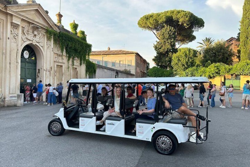 A white golf cart with passengers, parked in front of a historic building with an arched entrance and ivy. 1