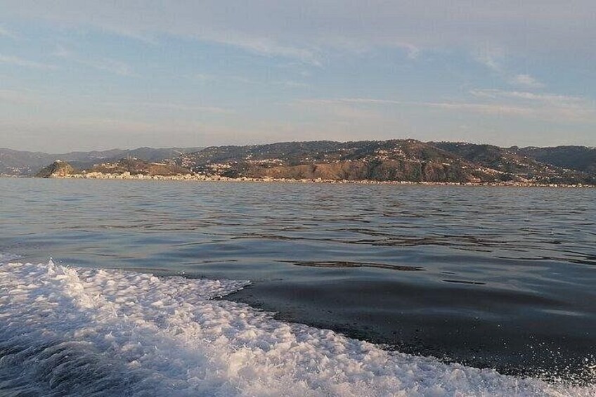 A calm body of water with waves breaking on the shore, a distant shoreline with buildings, and a hilly landscape under a clear sky. 4