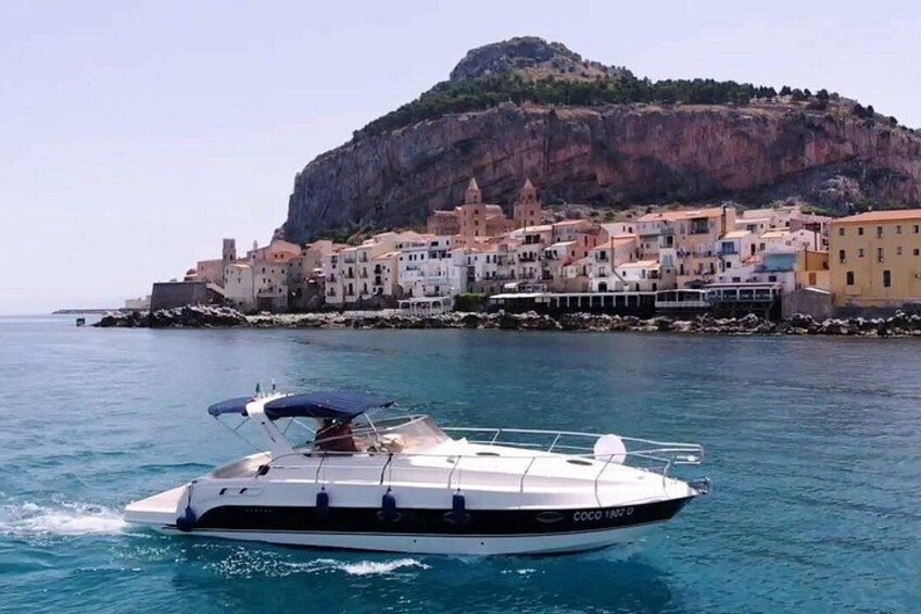 A white speedboat with a blue canopy on calm waters, a coastal town with colorful buildings, and a prominent cliff in the background. 2