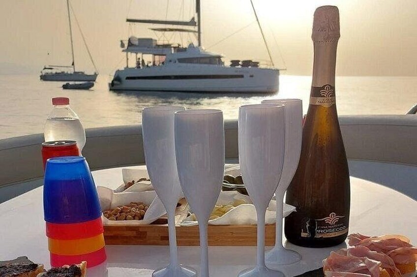 A table with a bottle of champagne, glasses, a bottle of water, and a plate of food on a boat with a sunset in the background. 3