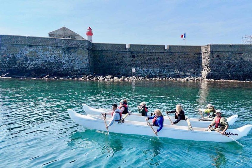 A group of people rowing a white boat near a stone wall with a lighthouse in the background. 4