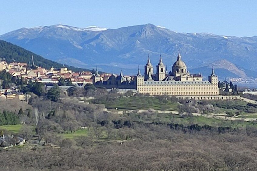 A large historical building with multiple domes, surrounded by a town and mountains in the background. 1