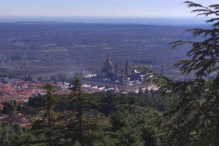 A cathedral with multiple spires, surrounded by a town with red-roofed buildings, set against a backdrop of a forested landscape and distant hills. 4