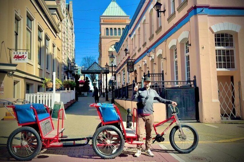 A person standing next to a red bicycle with a sidecar, a building with a green dome, and a street with buildings on either side. 1