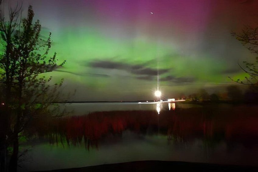 A nighttime landscape with a body of water reflecting lights, a tree, and the northern lights. 3