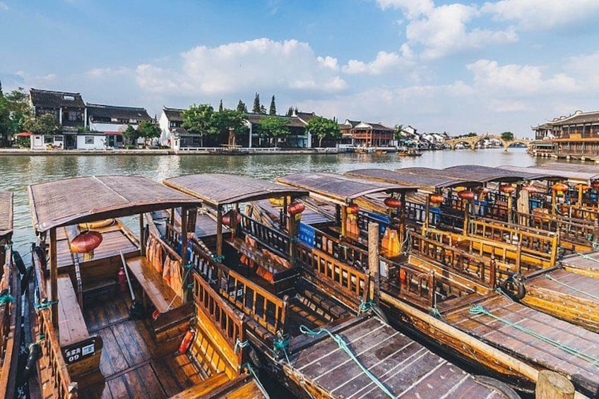 Ein traditionelles Holzboot mit einem Reetdach, Holzbänken und einem kleinen Tisch, das an einem Flussufer vor Gebäuden und einer Brücke liegt. 5
