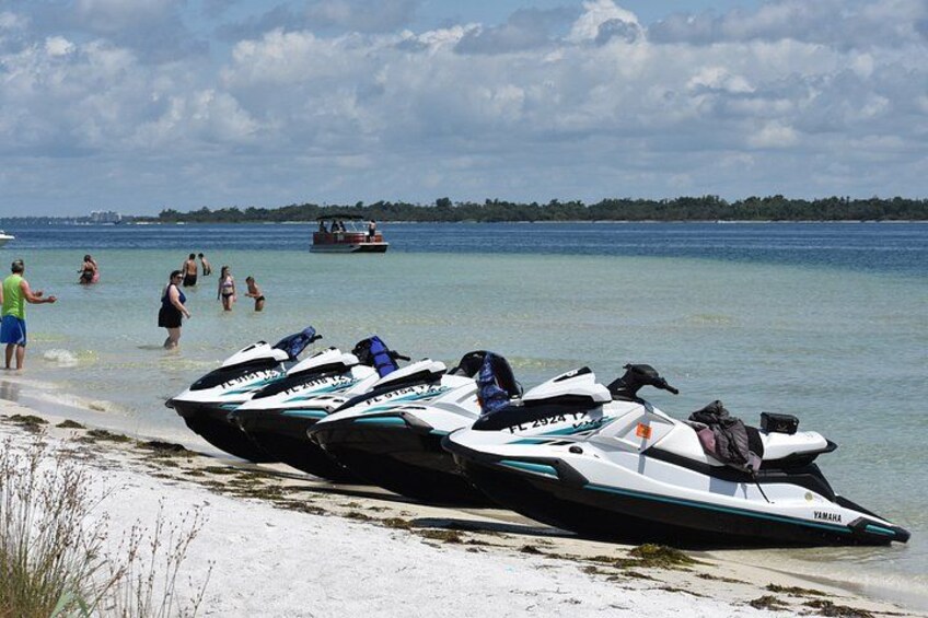 Motos acuáticas alineadas en una playa arenosa, con gente en el agua y un barco al fondo. 4