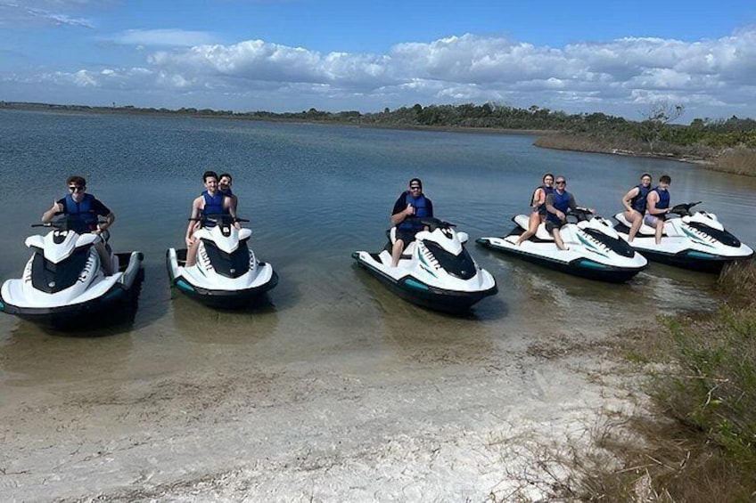 Fem personer på vandscootere på et roligt vandareal med en sandstrand og en klar himmel. 2