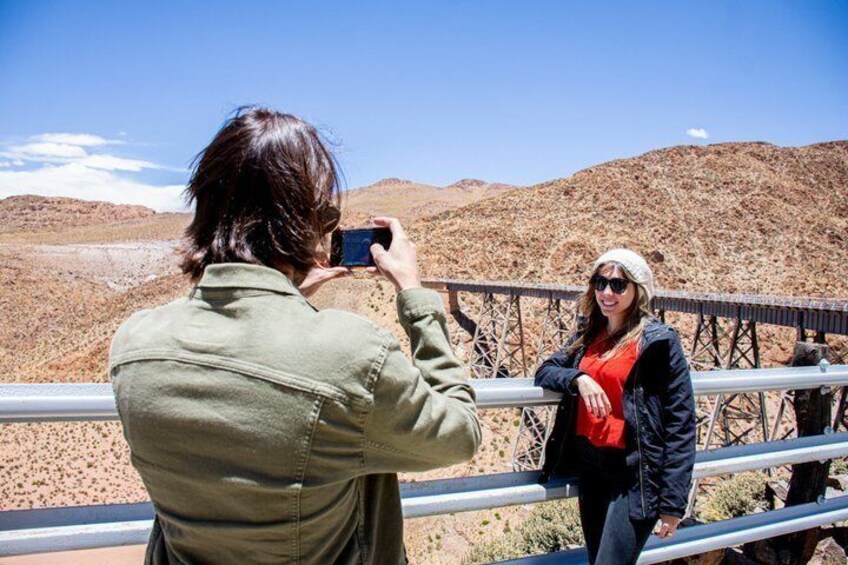 A person taking a photo of another person standing on a bridge with a desert landscape in the background. 3