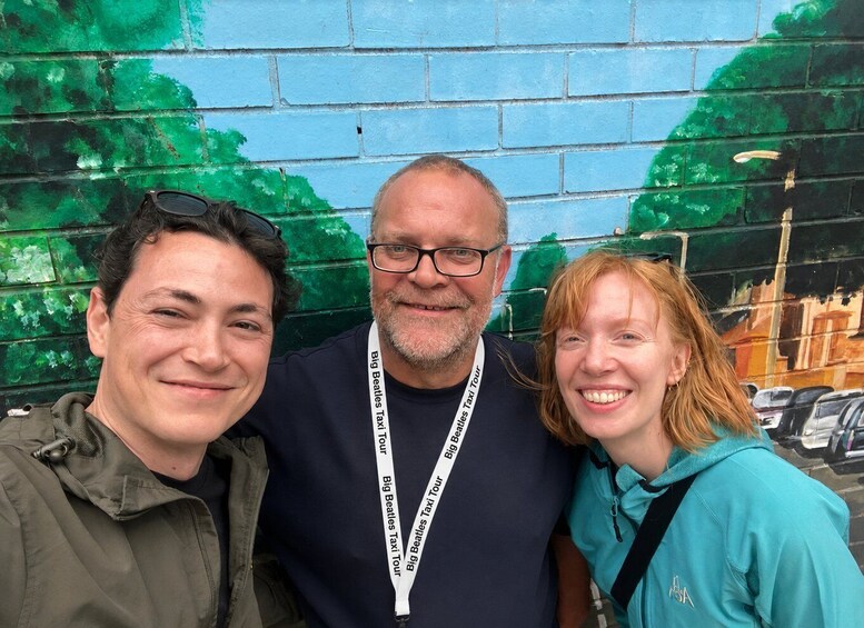 Three people posing in front of a mural with a blue brick wall and green foliage. 5