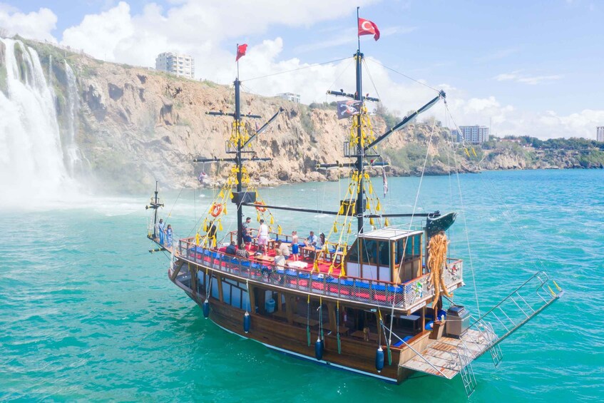 A boat with multiple flags, passengers, and a waterfall in the background. 1