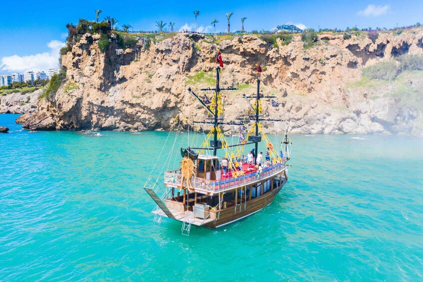 A wooden boat with passengers, anchored near a rocky coastline with buildings in the background. 2