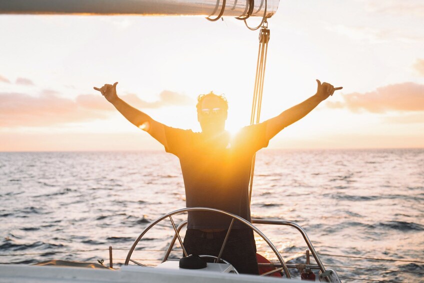 Eine Person auf einem Segelboot, die mit erhobenen Armen dem Sonnenuntergang entgegenblickt. 1